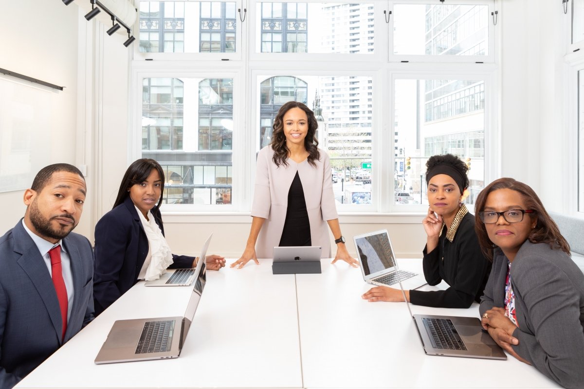 Five people in business attire sit around a conference table with laptops open; one woman stands at the head of the table beside a tablet, in front of floor-to-ceiling windows overlooking a city skyline.